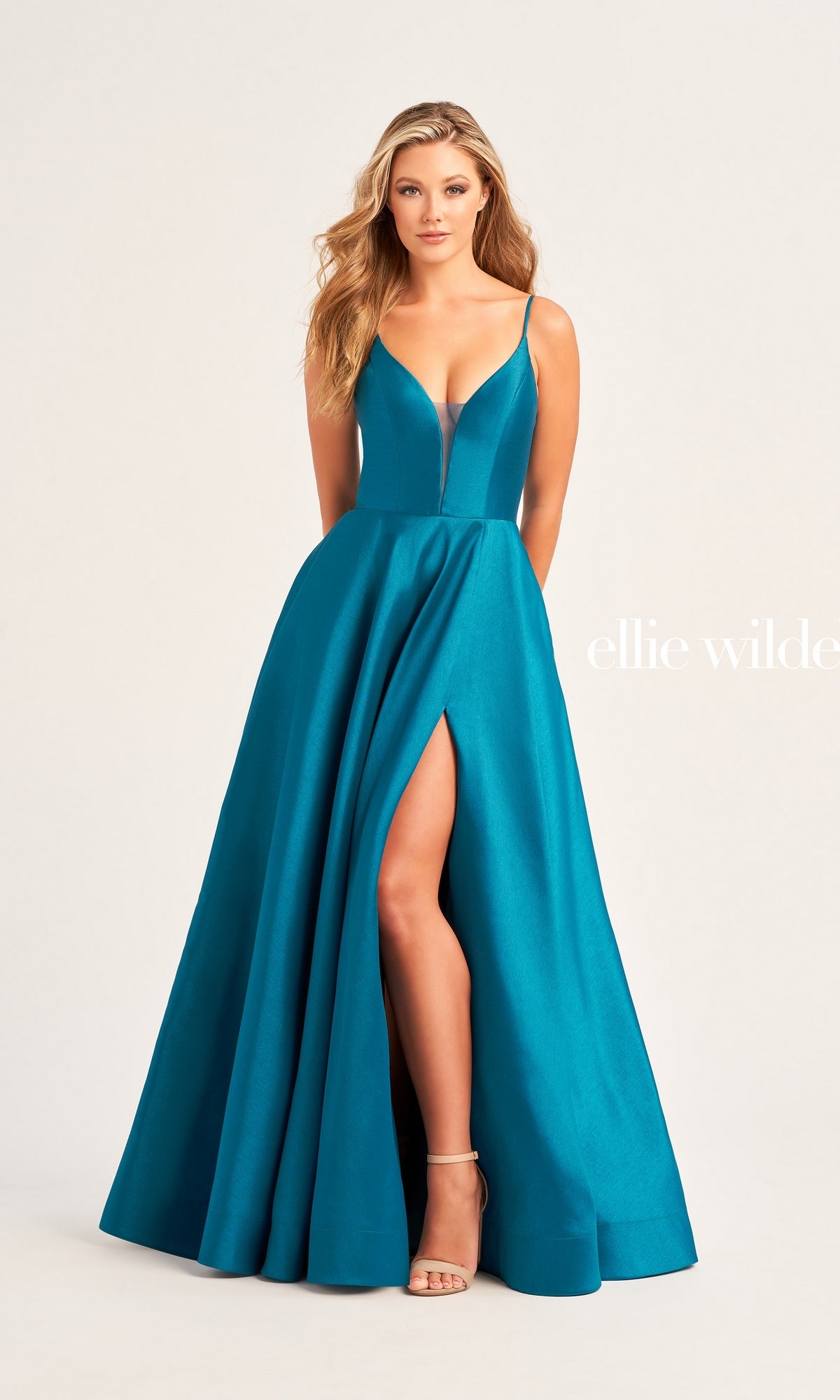 A woman models the Ellie Wilde Long A-Line Satin Prom Dress EW35232 in teal, featuring a deep V-neckline, thin strappy back, and high front slit, styled with nude heels.